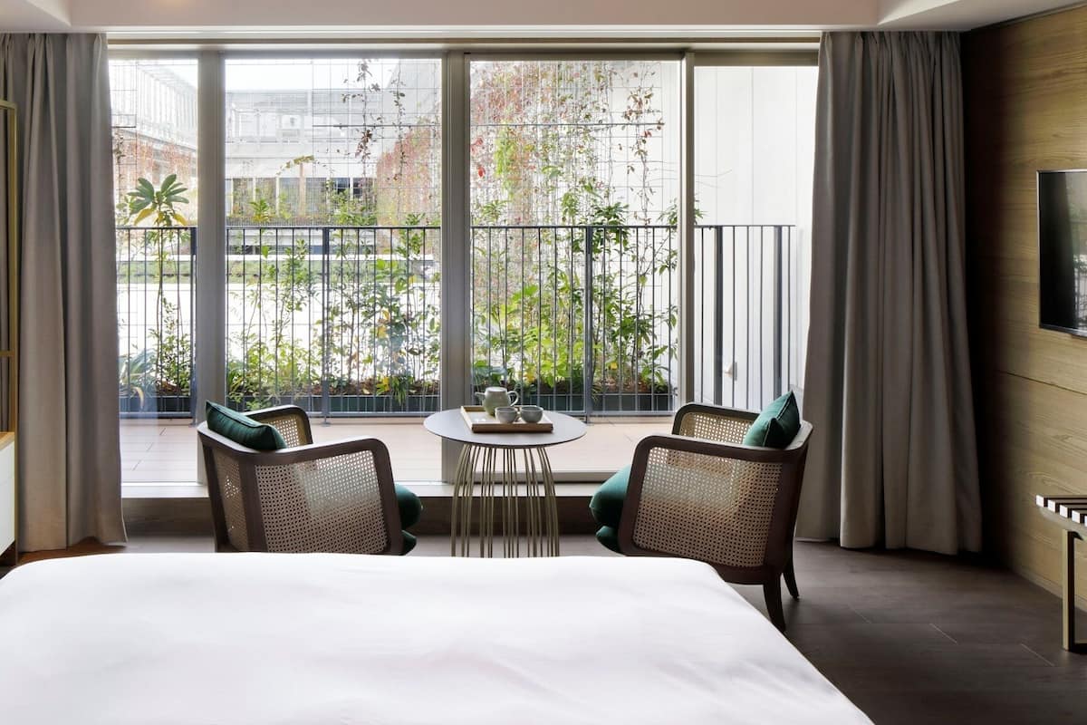 Et hotelværelse med en stor seng, to stole, et lille bord, et fjernsyn og en balkon med planter.