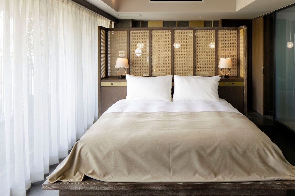 A hotel room with a large bed, two bedside lamps, a wooden headboard, and a glass partition.