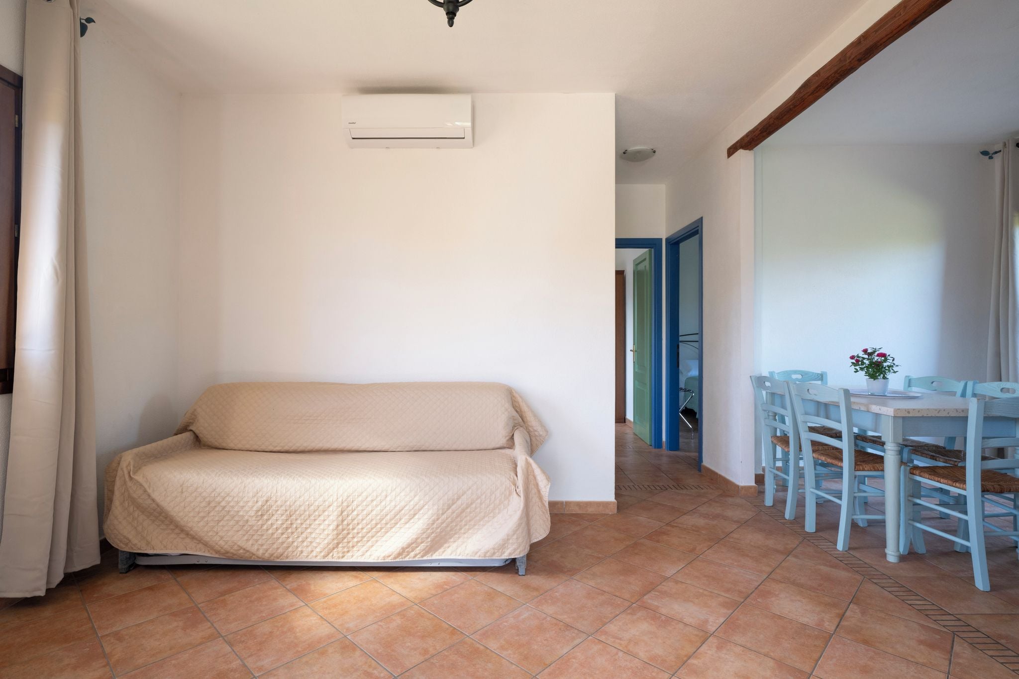 A room with a sofa, a dining table with chairs, an air conditioning unit, and a door leading to another room.