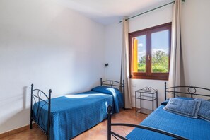 A room with two single beds, a window with curtains, a small table, and a view of greenery outside.