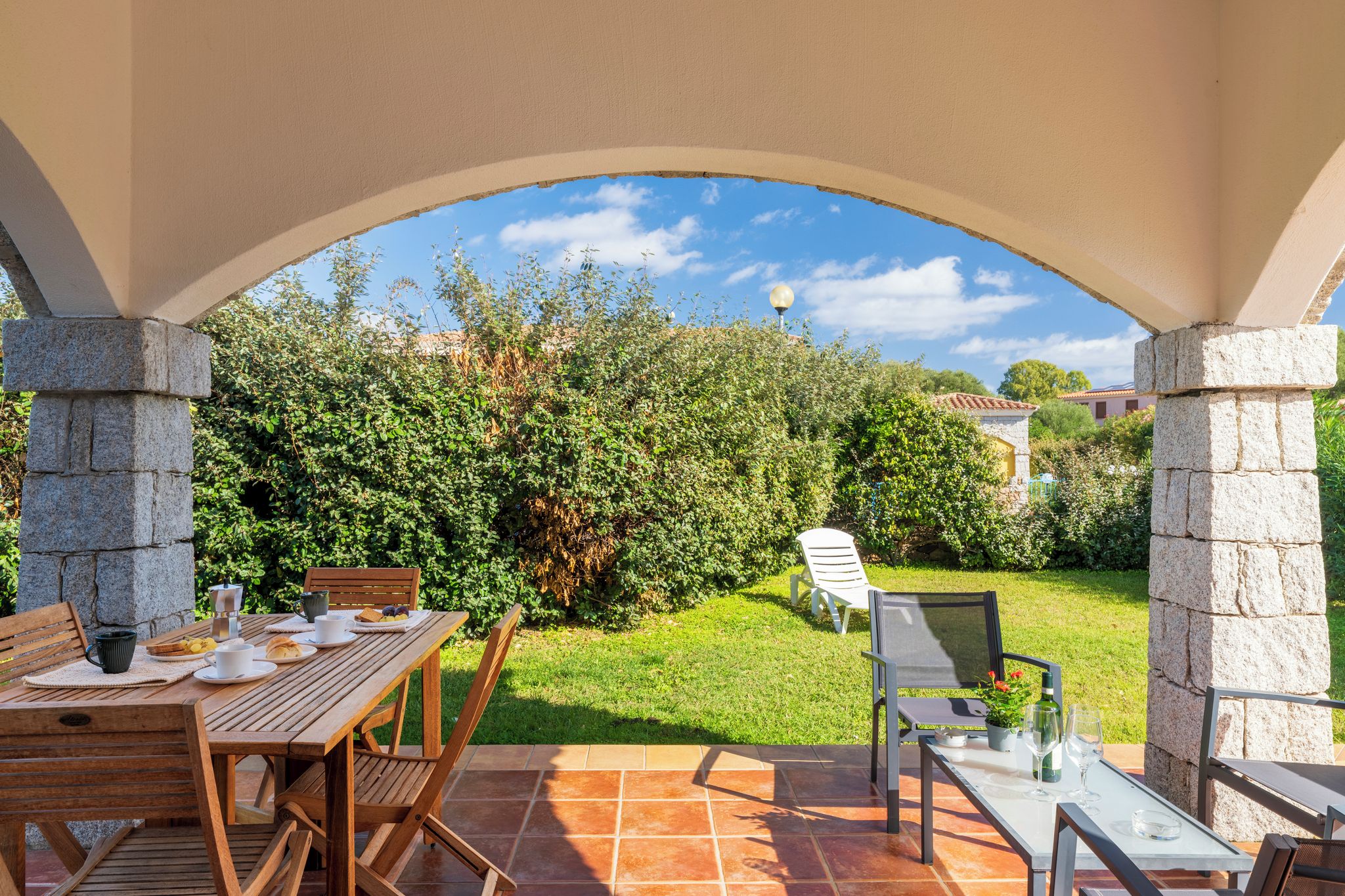 Un patio al aire libre con una mesa dispuesta para comer, rodeado de pilares de piedra y vistas a un jardín exuberante.