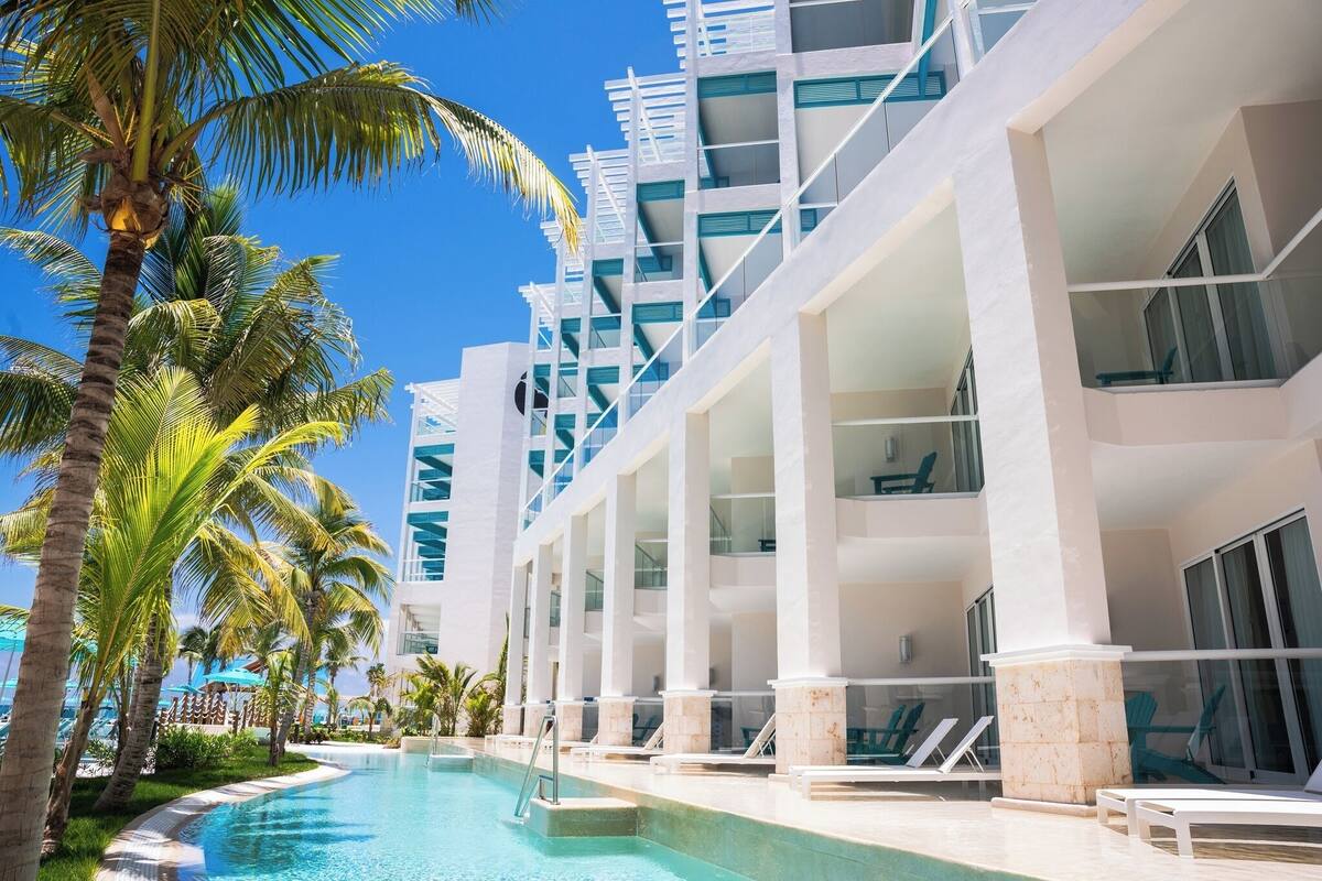 Un hotel moderno con piscina, palmeras y un cielo azul despejado.