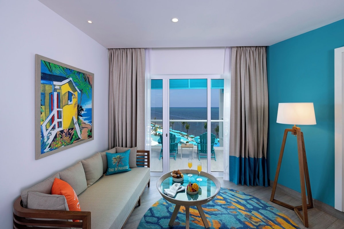 A hotel room with a sofa, a glass table with breakfast, a view of the ocean, and a painting on the wall.