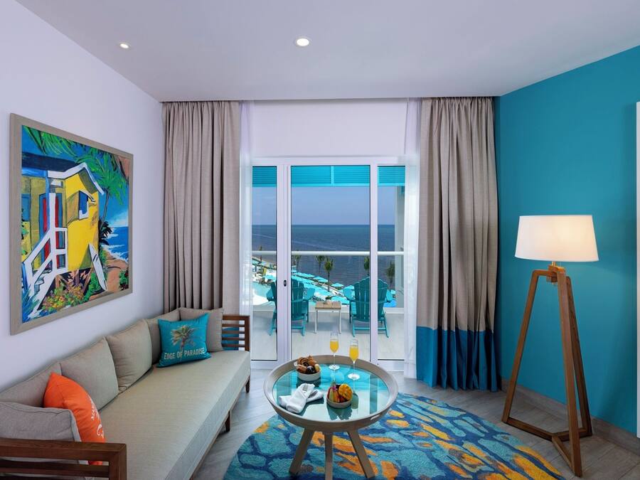 Habitación de hotel con sofá, una mesa de vidrio con desayuno, vista al océano y un cuadro en la pared.