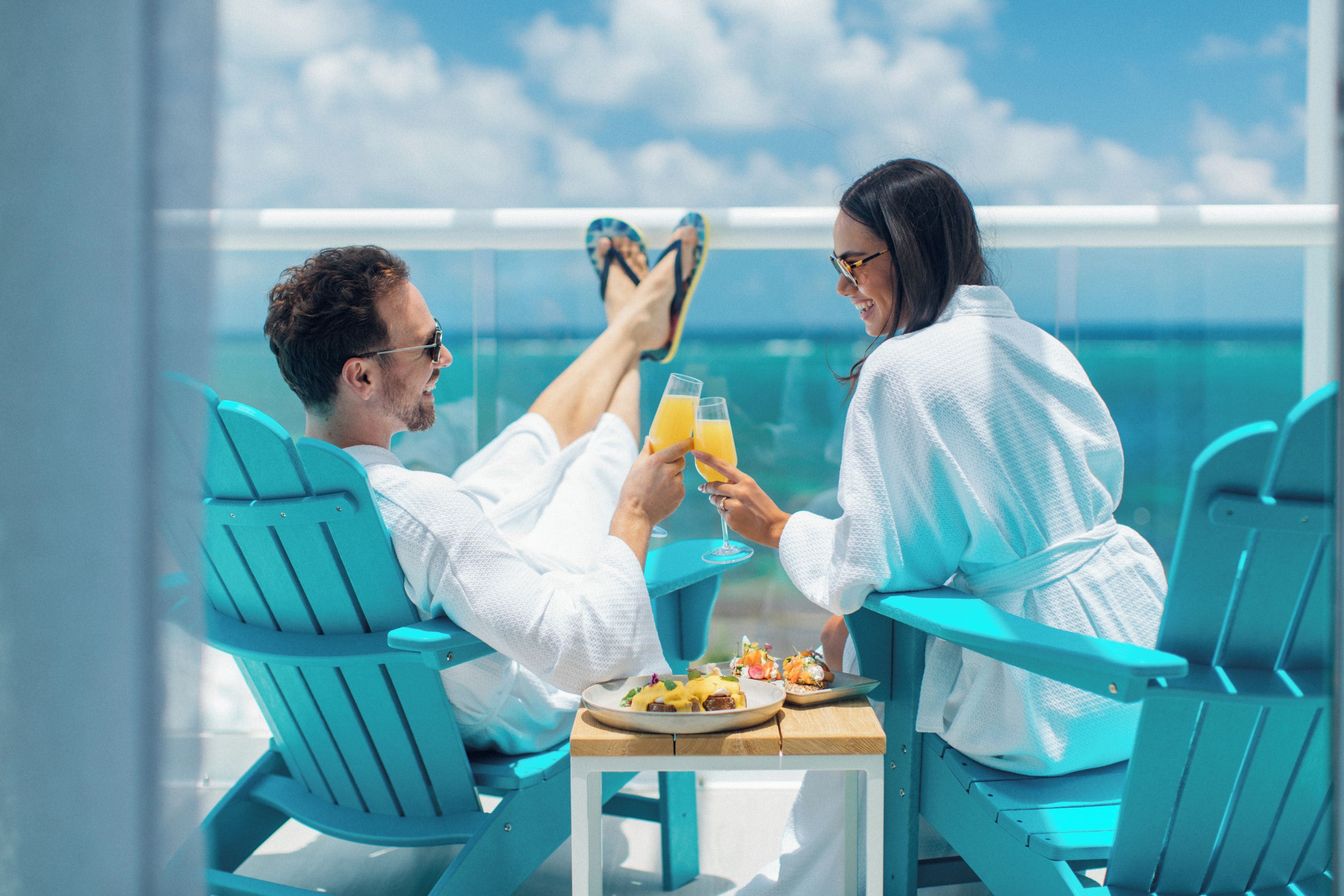 Deux personnes vêtues de longues robes blanches sont assises sur des chaises turquoise, autour d’une table où se trouvent des boissons et de la nourriture. Elles ont devant elles une étendue d’eau.