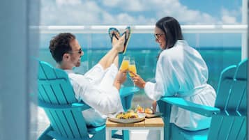 Deux personnes vêtues de longues robes blanches sont assises sur des chaises turquoise, autour d’une table où se trouvent des boissons et de la nourriture. Elles ont devant elles une étendue d’eau.