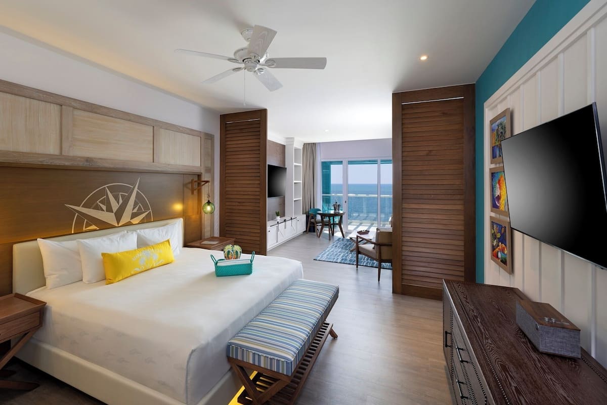 Habitación de hotel con una cama grande, ventilador de techo, televisor y vista al océano.