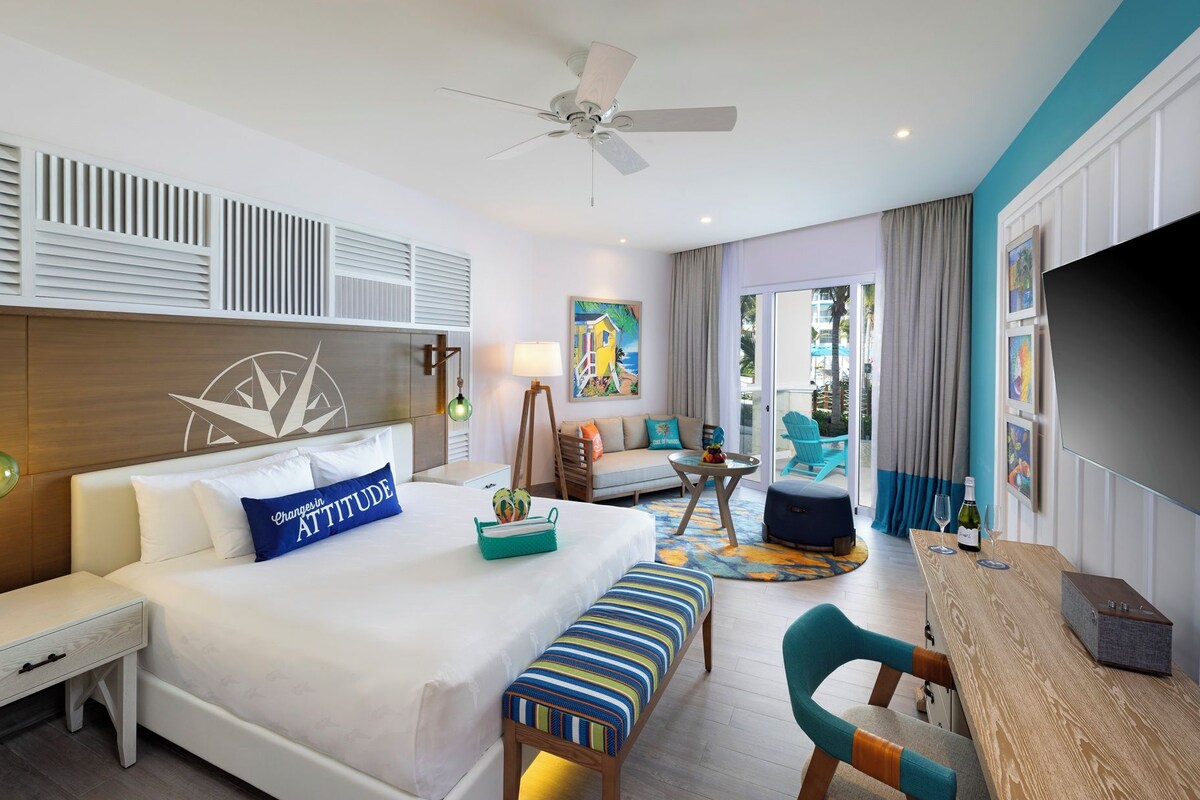 A hotel room with a bed, a desk, a chair, a TV, and a balcony with a view of the outside.
