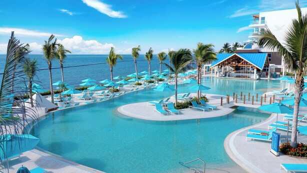 Piscina de un resort con agua azul cristalina, rodeada de palmeras y sillas de descanso.