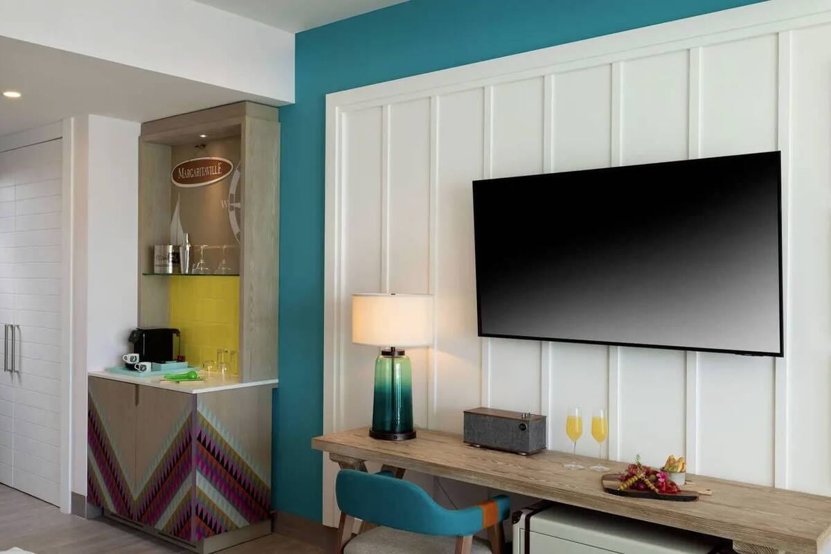Habitación de hotel con kitchenette, un televisor de pantalla plana en la pared, un escritorio con lámpara y una silla.