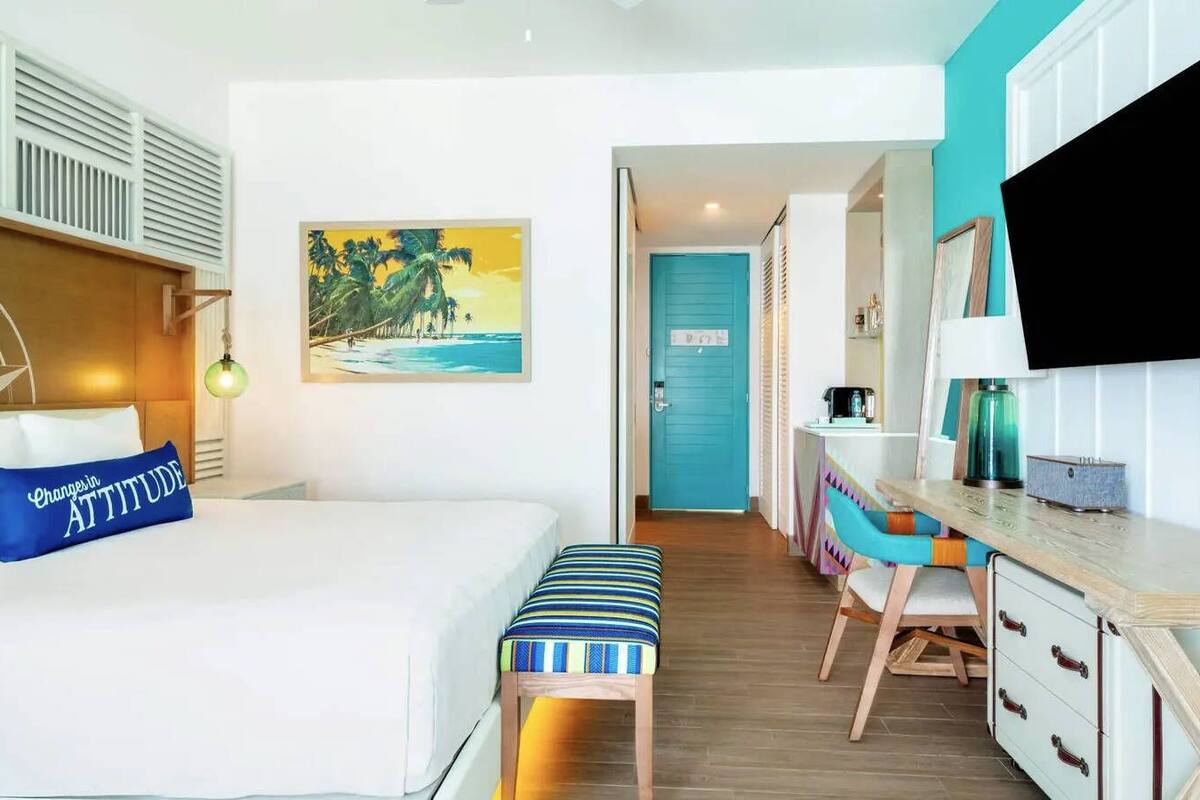 Una habitación de hotel con cama, escritorio, silla, televisor y un cuadro con una playa.