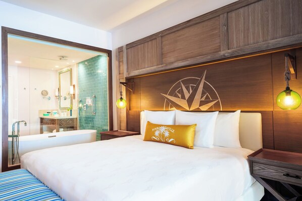 A hotel room with a large bed, a wooden headboard, a nightstand, a bedside table, a mirror, and a bathroom with a bathtub and a sink. - Margaritaville Island Reserve Riviera Maya (Puerto Morelos)