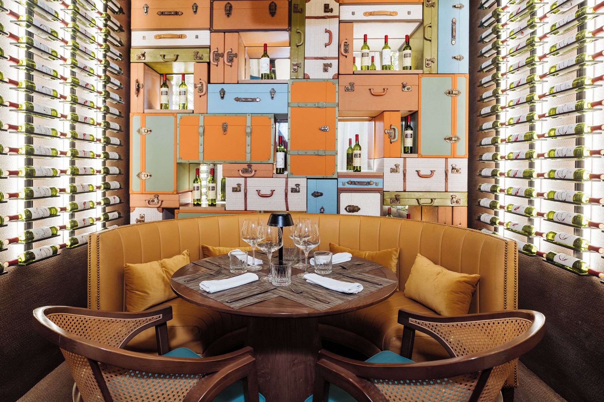 A dining area with a round table surrounded by wicker chairs, set against a wall of vintage suitcases filled with wine bottles.