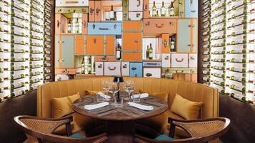 A dining area with a round table surrounded by wicker chairs, set against a wall of vintage suitcases filled with wine bottles.