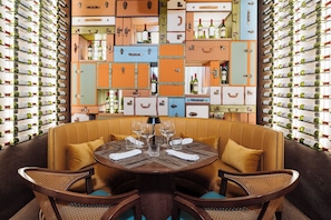 A dining area with a round table surrounded by wicker chairs, set against a wall of vintage suitcases filled with wine bottles. - Margaritaville Island Reserve Riviera Maya (Puerto Morelos)