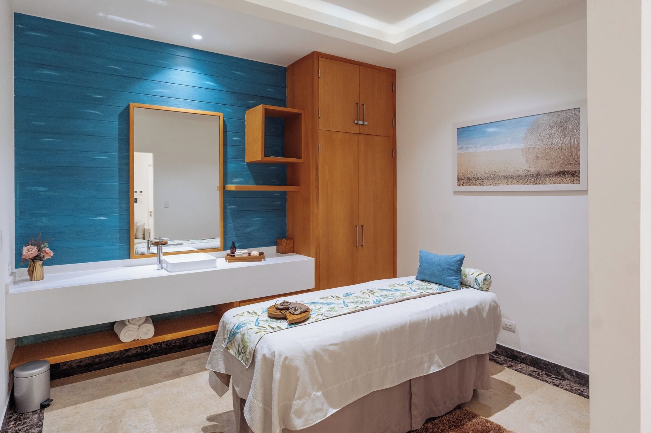 A modern bathroom with a large mirror, wooden cabinets, a white vanity, and a bed with a blue pillow.