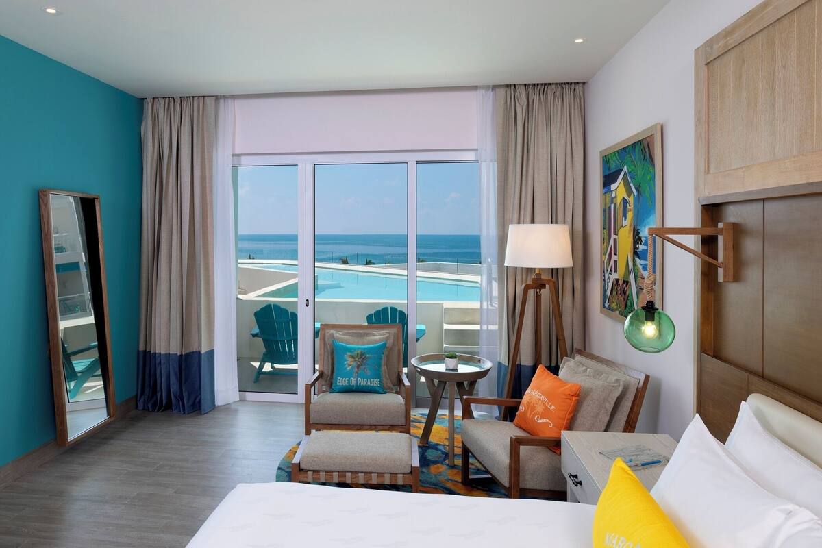 Habitación de hotel con un ventanal que ofrece vistas al océano, cama con ropa blanca, sillón con cojín, mesita con lámpara y un cuadro en la pared.