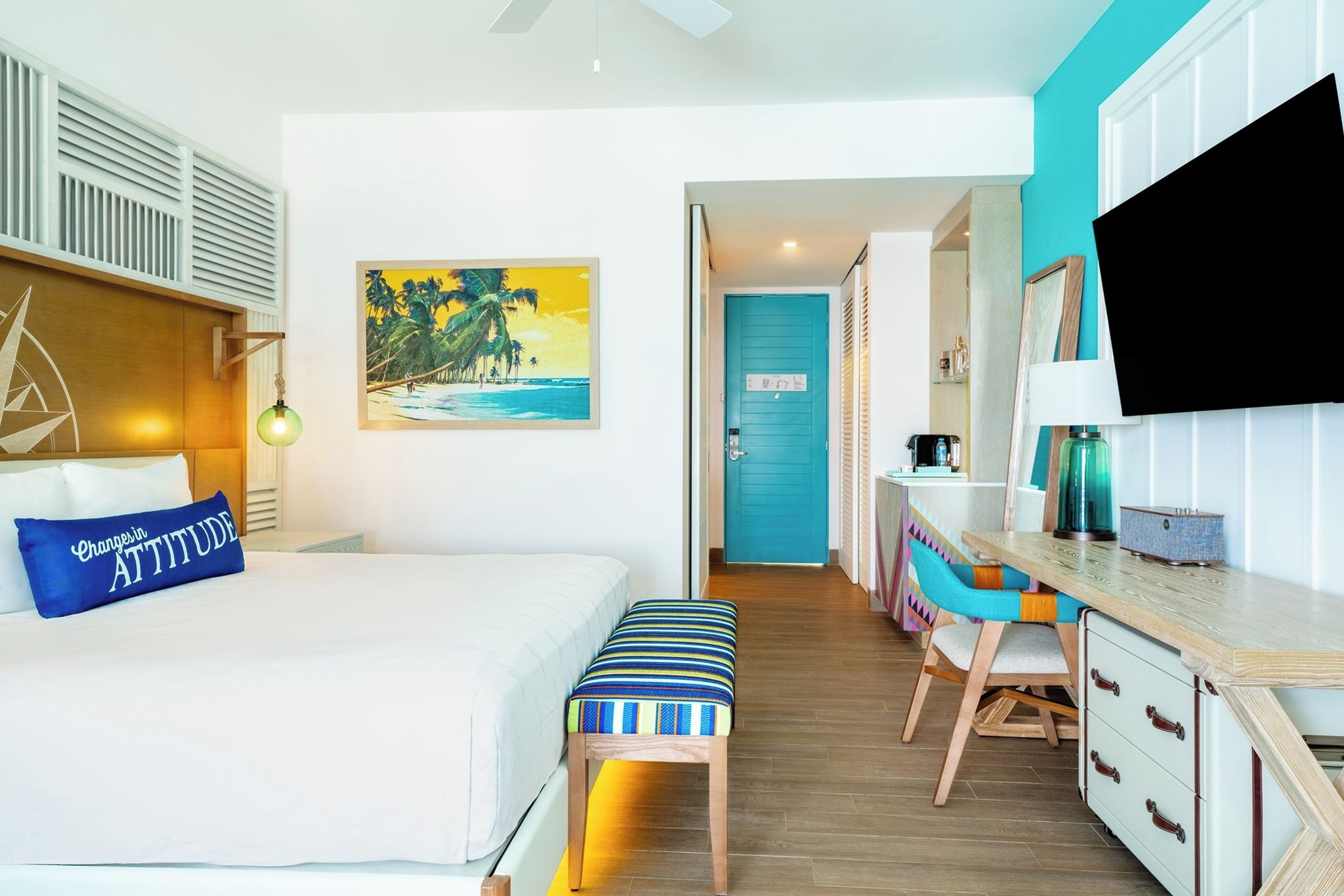 A hotel room with a bed, a desk, a chair, a TV, and a painting of a beach scene.