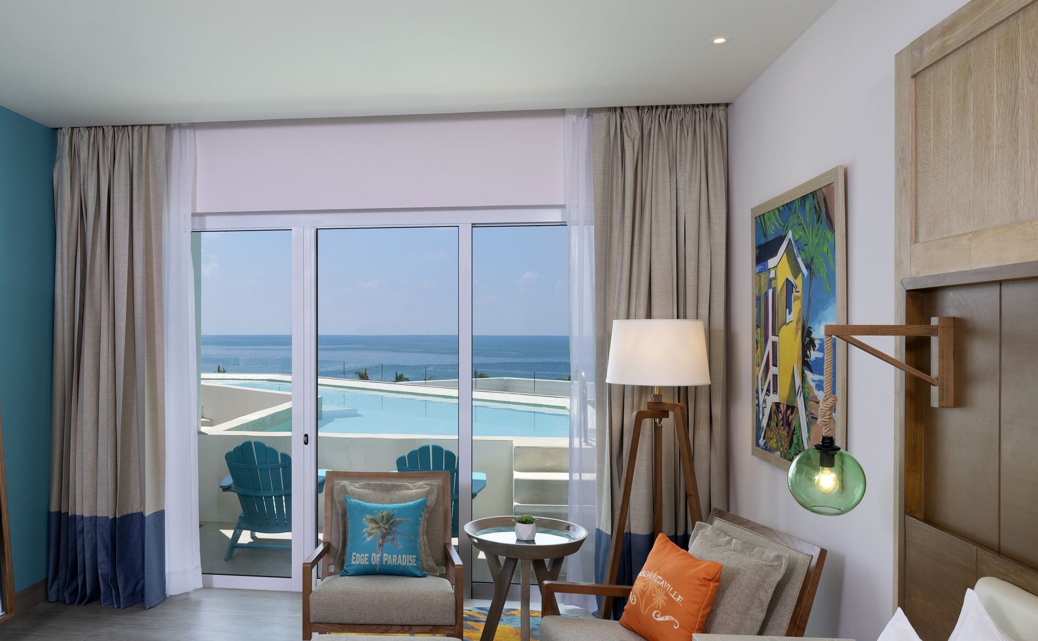 A hotel room with a large sliding glass door leading to a pool area, a chair with a cushion, a small table with a plant, a lamp, and a painting on the wall.