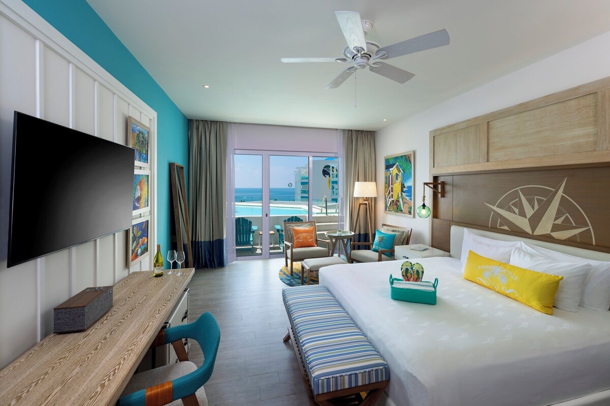 A hotel room with a bed, a desk, a TV, a ceiling fan, and a view of the ocean.