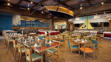 Une grande salle à manger avec de nombreuses tables et chaises, un coin bar et un présentoir de cannes à pêche accroché au mur.