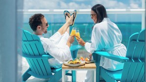 Dos personas vestidas de blanco sentadas en sillas turquesa junto a una mesa con bebidas y comida, con vista a una masa de agua.