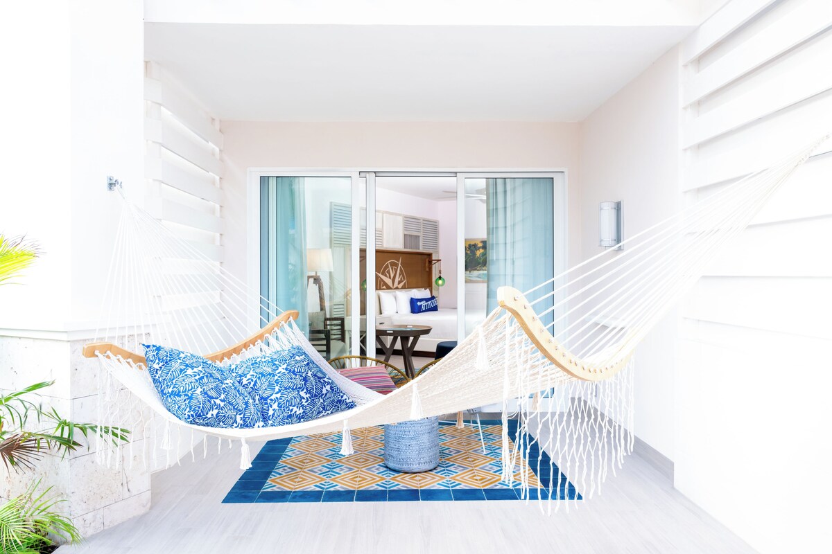 A hammock chair with blue patterned cushions in a bright room with a tiled floor and a view of a dining area through glass doors.