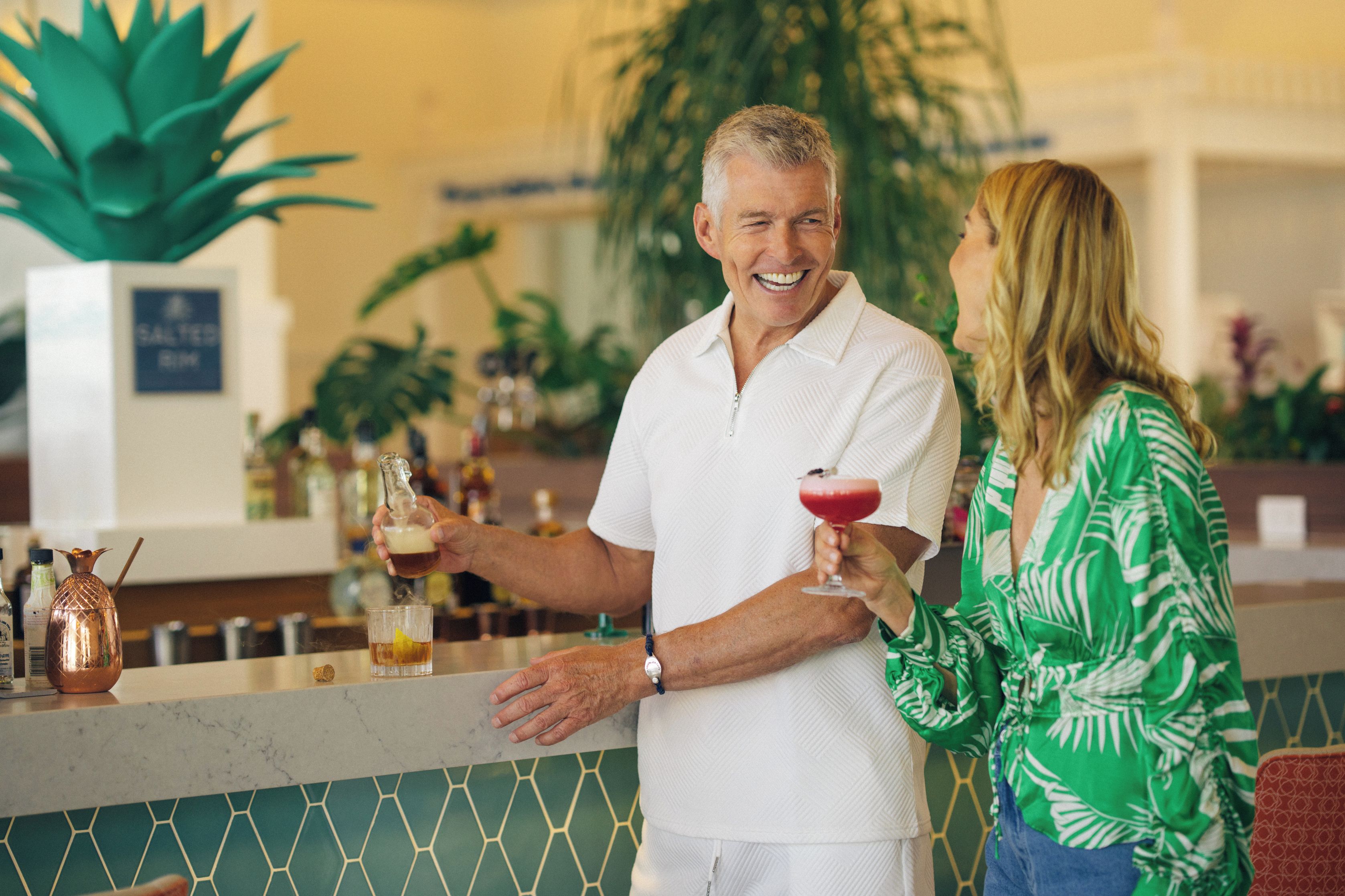 Un homme et une femme sont assis au bar, devant une carafe en cuivre et un verre contenant une boisson rouge.