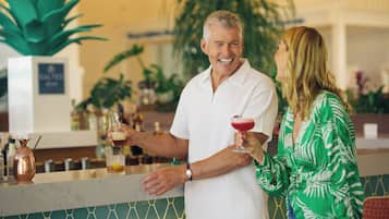 Un homme et une femme sont assis au bar, devant une carafe en cuivre et un verre contenant une boisson rouge.