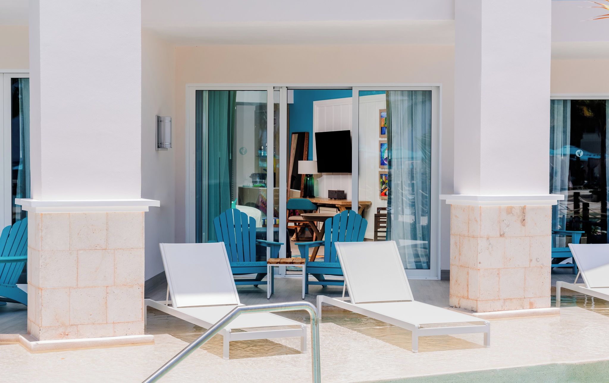 A poolside area with white lounge chairs, a blue chair, and a table with a lamp.