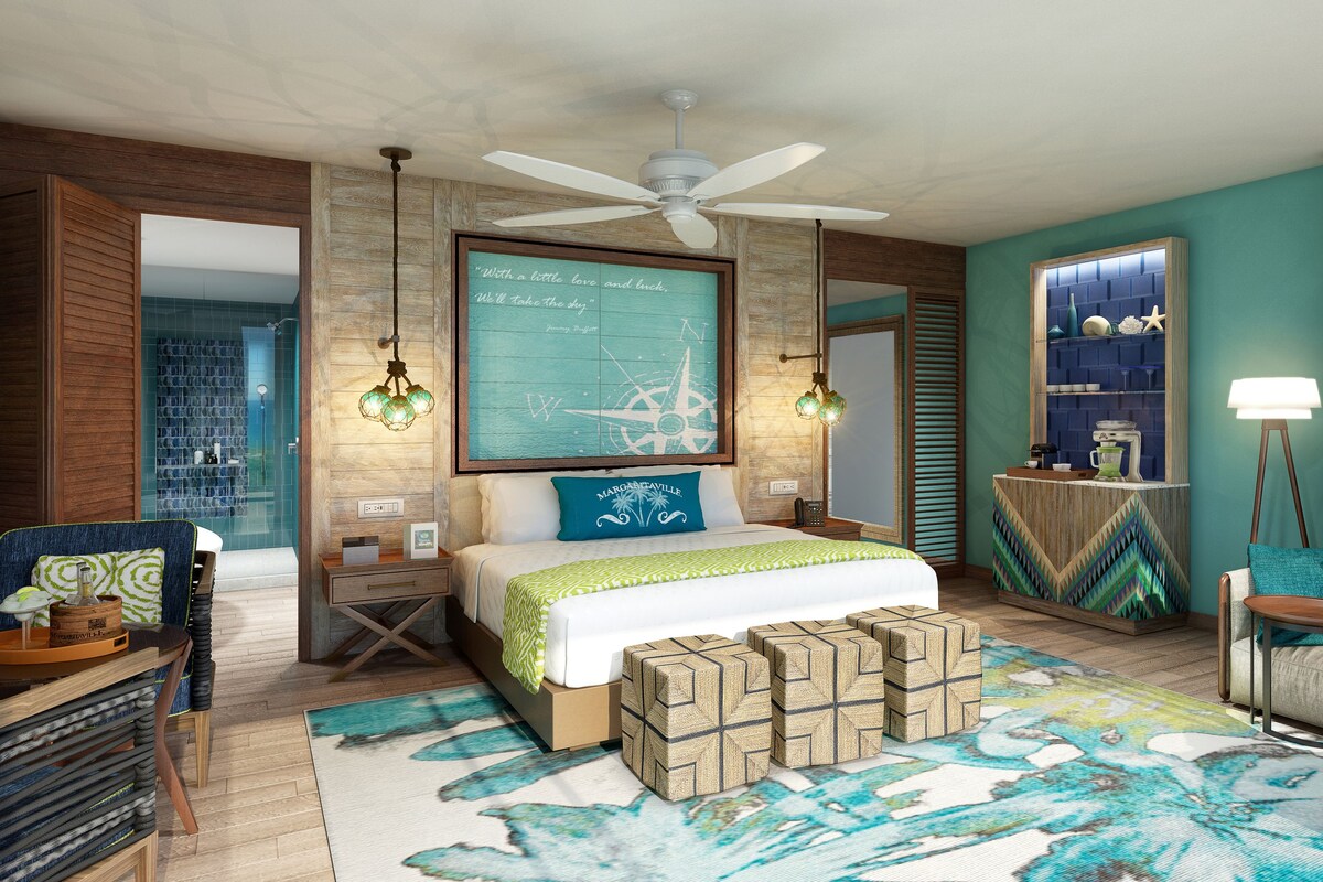 A modern hotel room with a large bed, a ceiling fan, a wall-mounted compass, and a view of the sea through a window.