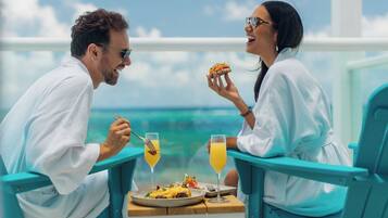 A couple dining at a table with a view of the ocean, a plate of food, and two drinks.
