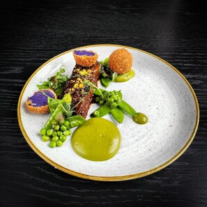 Un plato de comida con diversos ingredientes, incluyendo un trozo de carne, guisantes y una salsa verde.