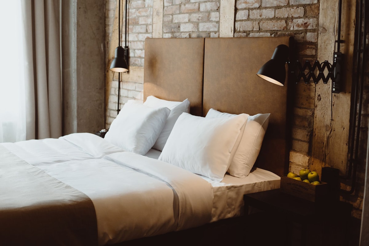 Una cama bien hecha con sábanas y almohadas blancas, un cabecero de madera y una mesita de noche con una lámpara y una caja de manzanas.