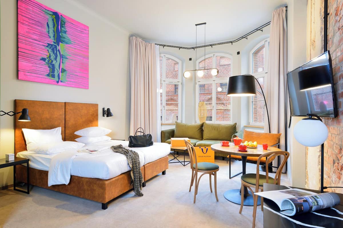A hotel room with a bed, a desk, a chair, a sofa, a TV, and a window with a view of a brick building.