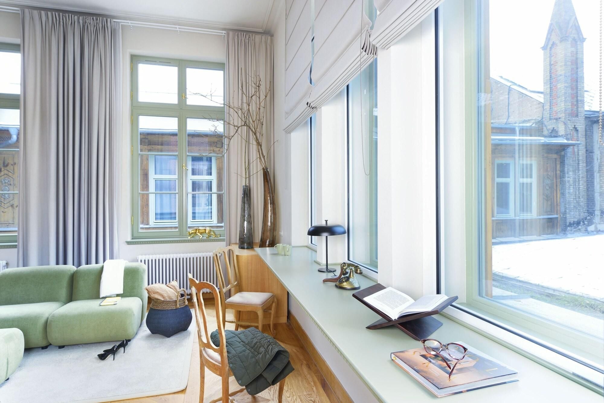 A modern living room with a green sofa, a wooden chair, a small table, and a large window offering a view of a building.