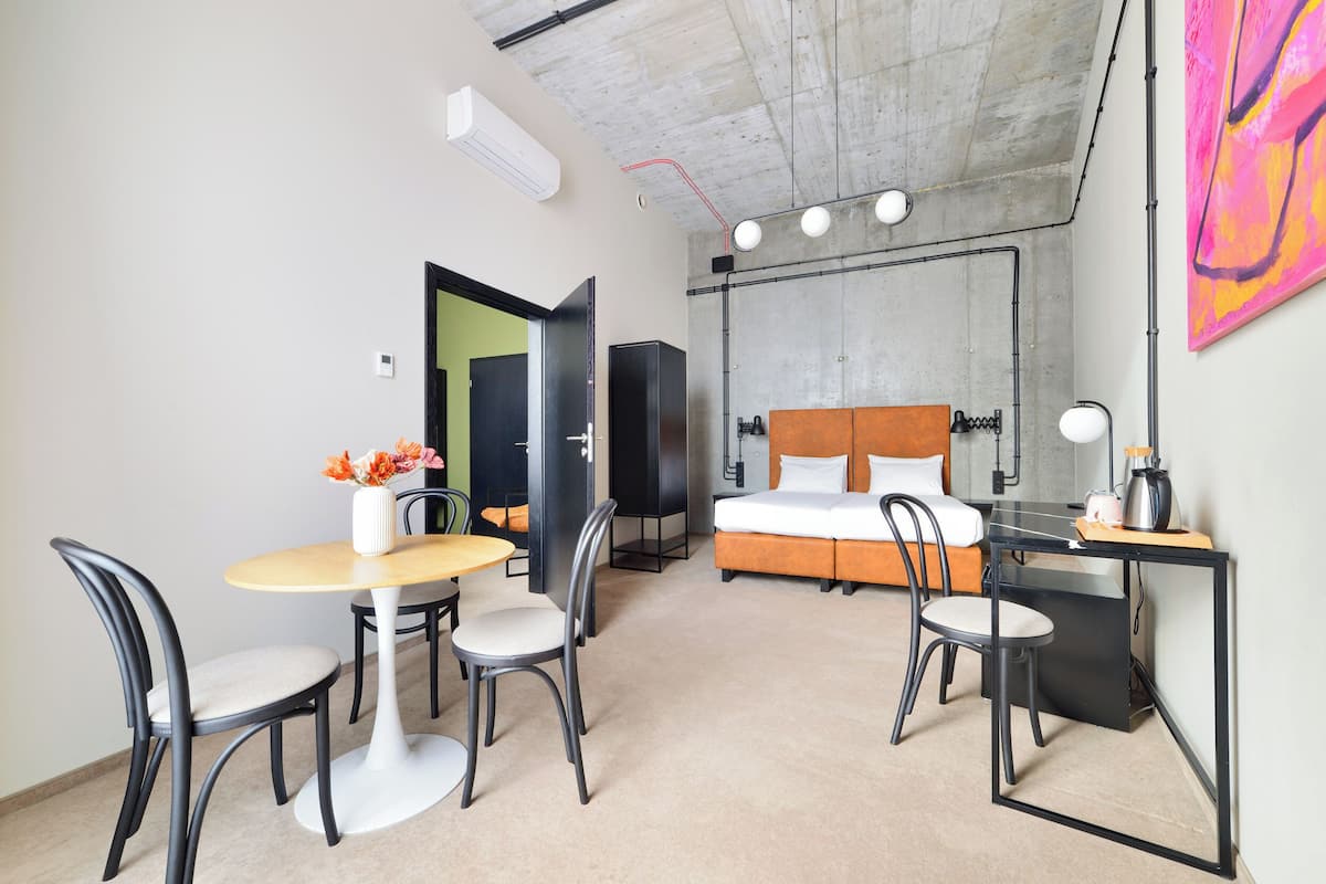 A modern hotel room with a bed, a desk, a chair, a table, and a small kitchenette.