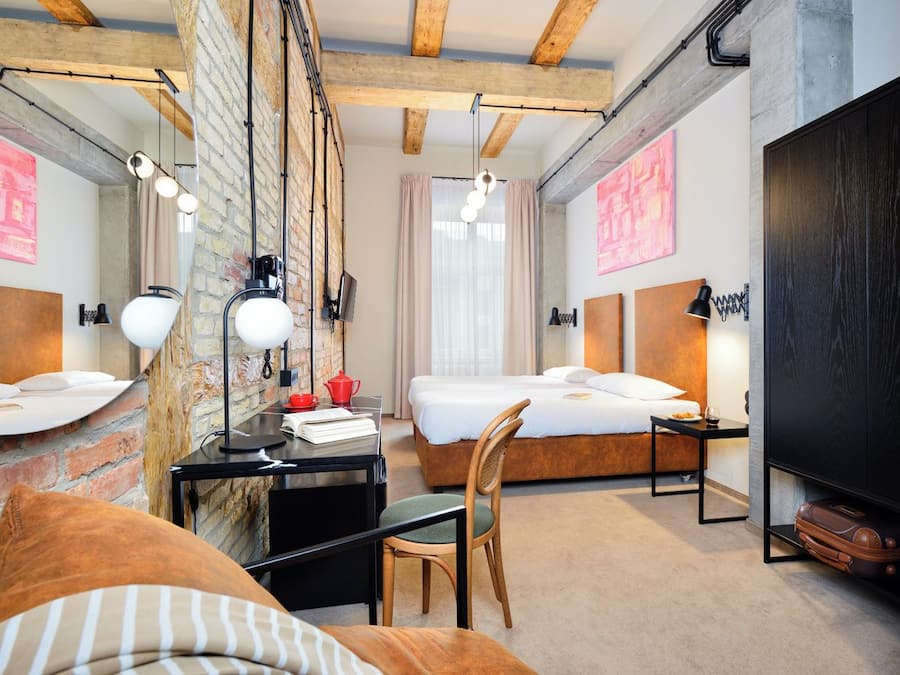 A modern hotel room with exposed brick walls, a loft bed, a desk with a chair, and a large wardrobe.