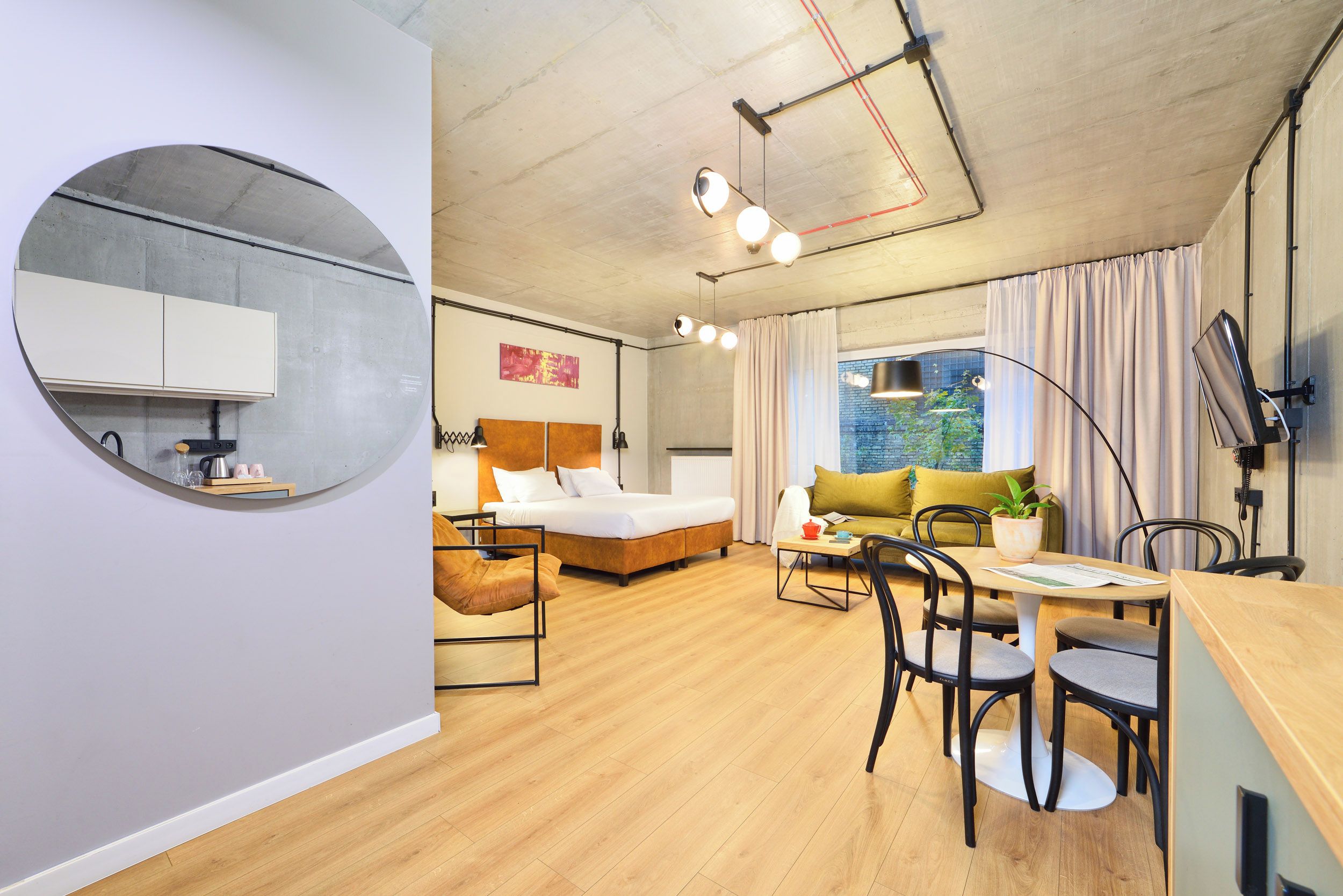 Een moderne hotelkamer met een bed, een bank, een eettafel met stoelen, een ronde spiegel en een keukenhoek.