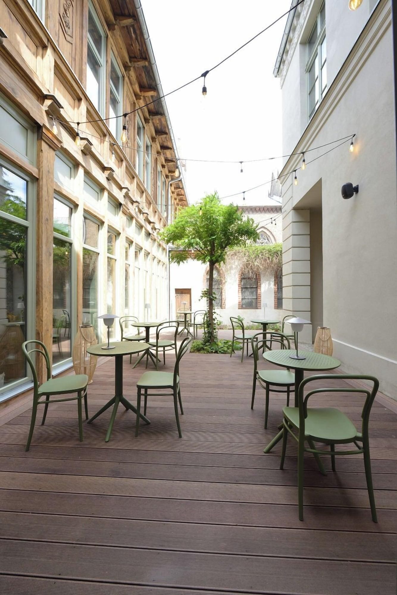 Ein Außenbereich mit Terrasse, Tischen und Stühlen, umgeben von Gebäuden mit großen Fenstern und einem Baum in der Mitte.