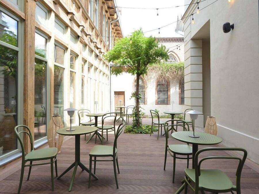Uma área de pátio ao ar livre com mesas e cadeiras, rodeada por edifícios com grandes janelas e uma árvore no centro.