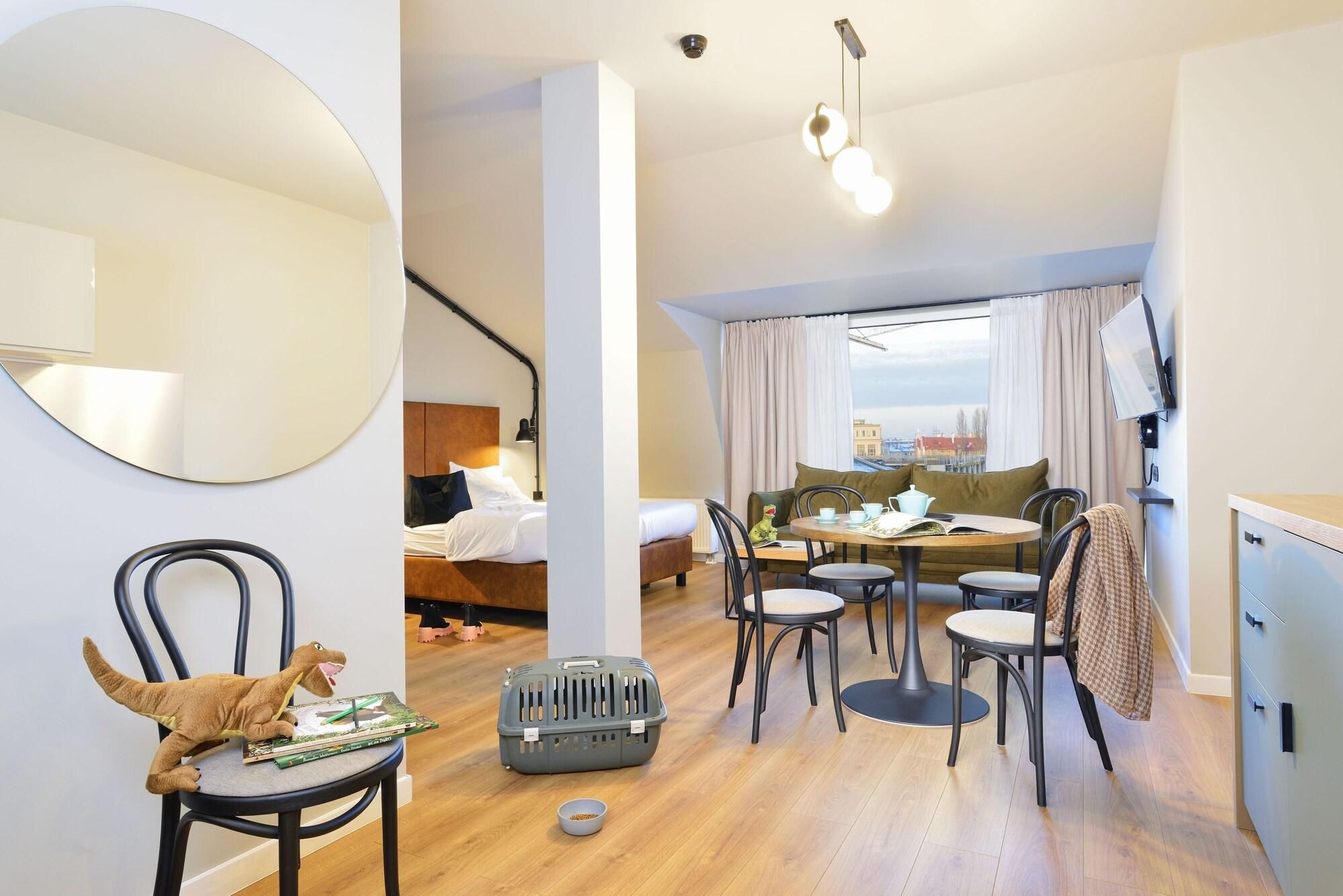 A modern hotel room with a bed, a dining area with a table and chairs, a large mirror, and a view of the city.