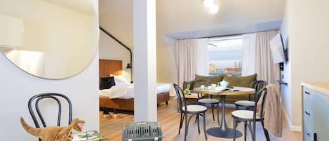 A modern hotel room with a bed, a dining area with a table and chairs, a large mirror, and a view of the city.