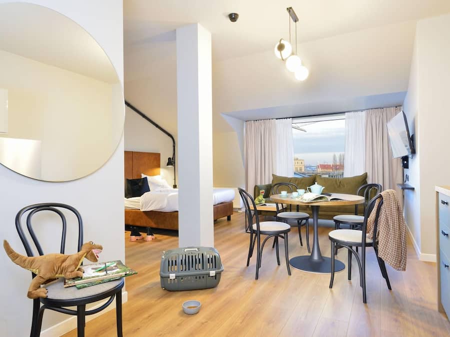 A modern hotel room with a bed, a dining area with a table and chairs, a large mirror, and a view of the city.