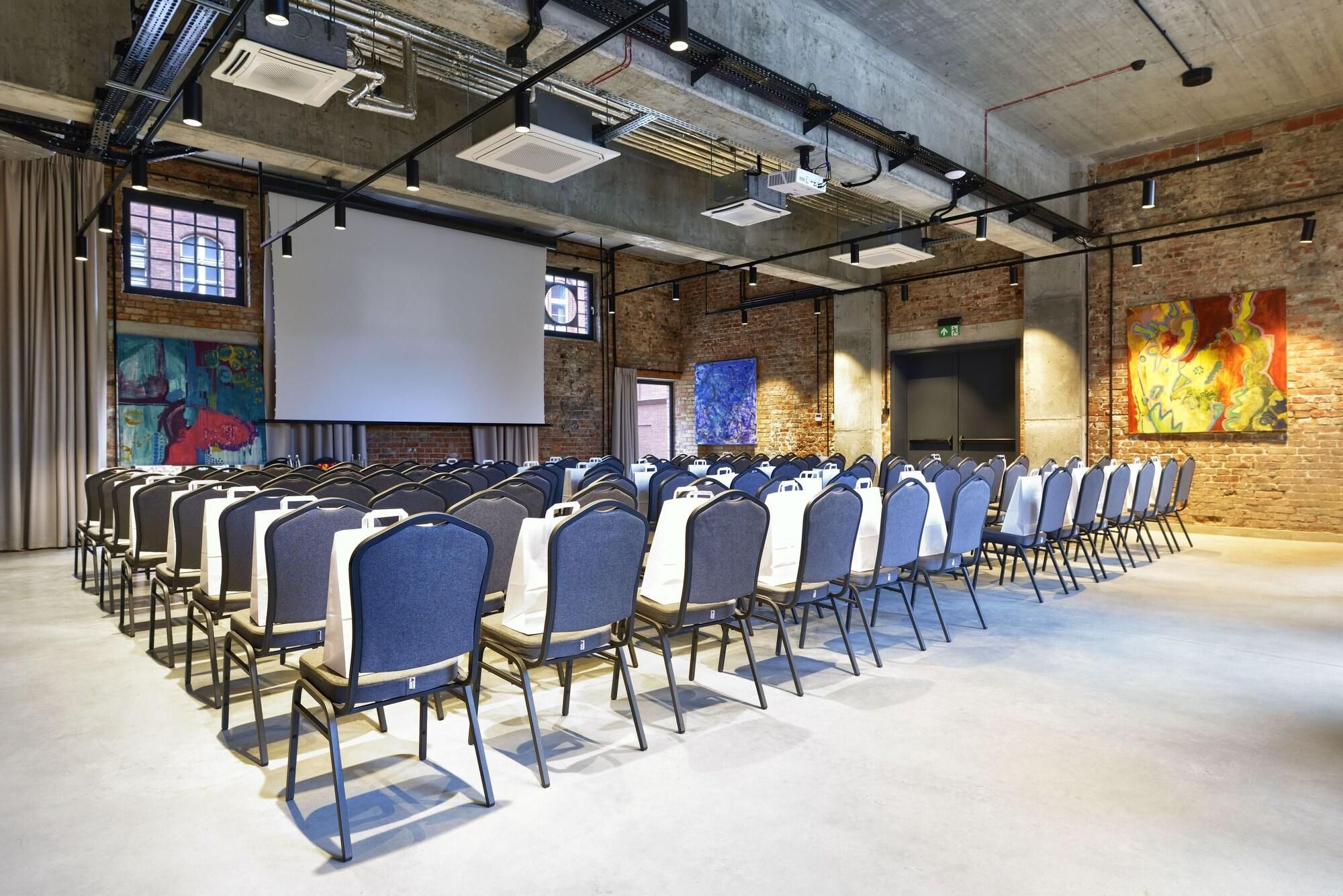 A conference room with rows of chairs, a projector screen, and a brick wall with artwork.