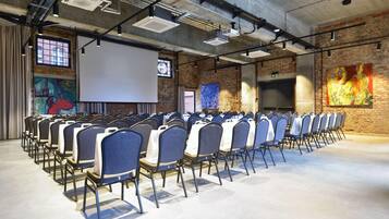 A conference room with rows of chairs, a projector screen, and a brick wall with artwork.