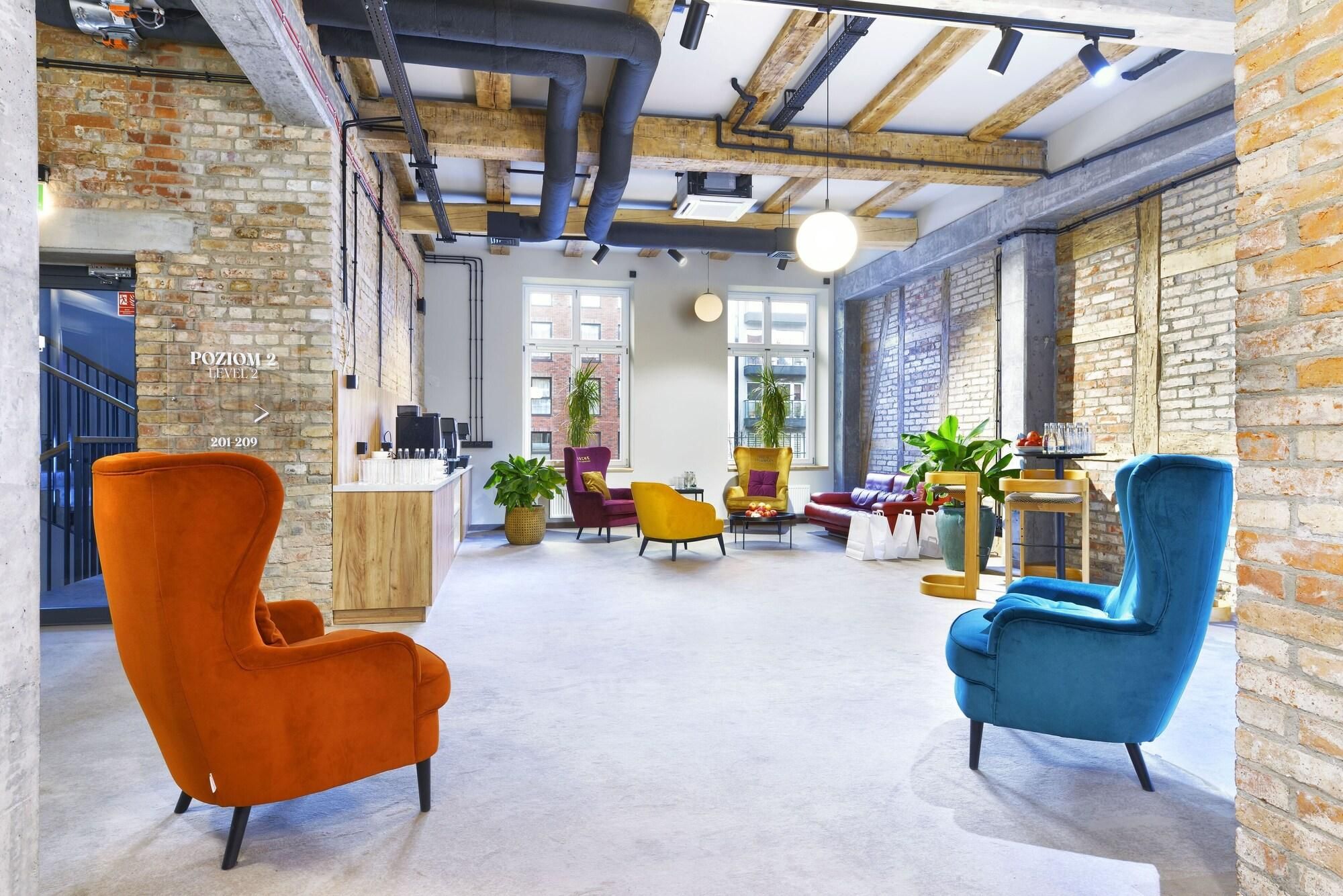 A spacious lobby with exposed brick walls, a high ceiling with visible beams, and a mix of seating including an orange armchair, a blue armchair, and a yellow armchair.