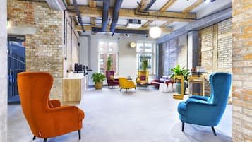 A spacious lobby with exposed brick walls, a high ceiling with visible beams, and a mix of seating including an orange armchair, a blue armchair, and a yellow armchair.