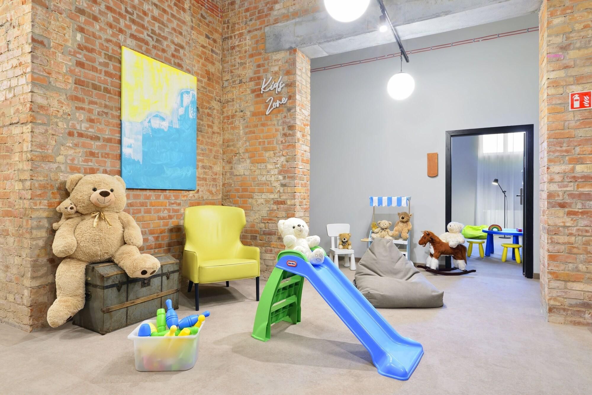Un área de juegos infantiles con pared de ladrillo, una silla amarilla, un osito de peluche, un tobogán y varios juguetes.