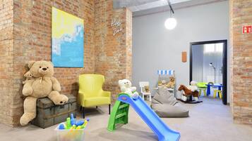 Un área de juegos infantiles con pared de ladrillo, una silla amarilla, un osito de peluche, un tobogán y varios juguetes.